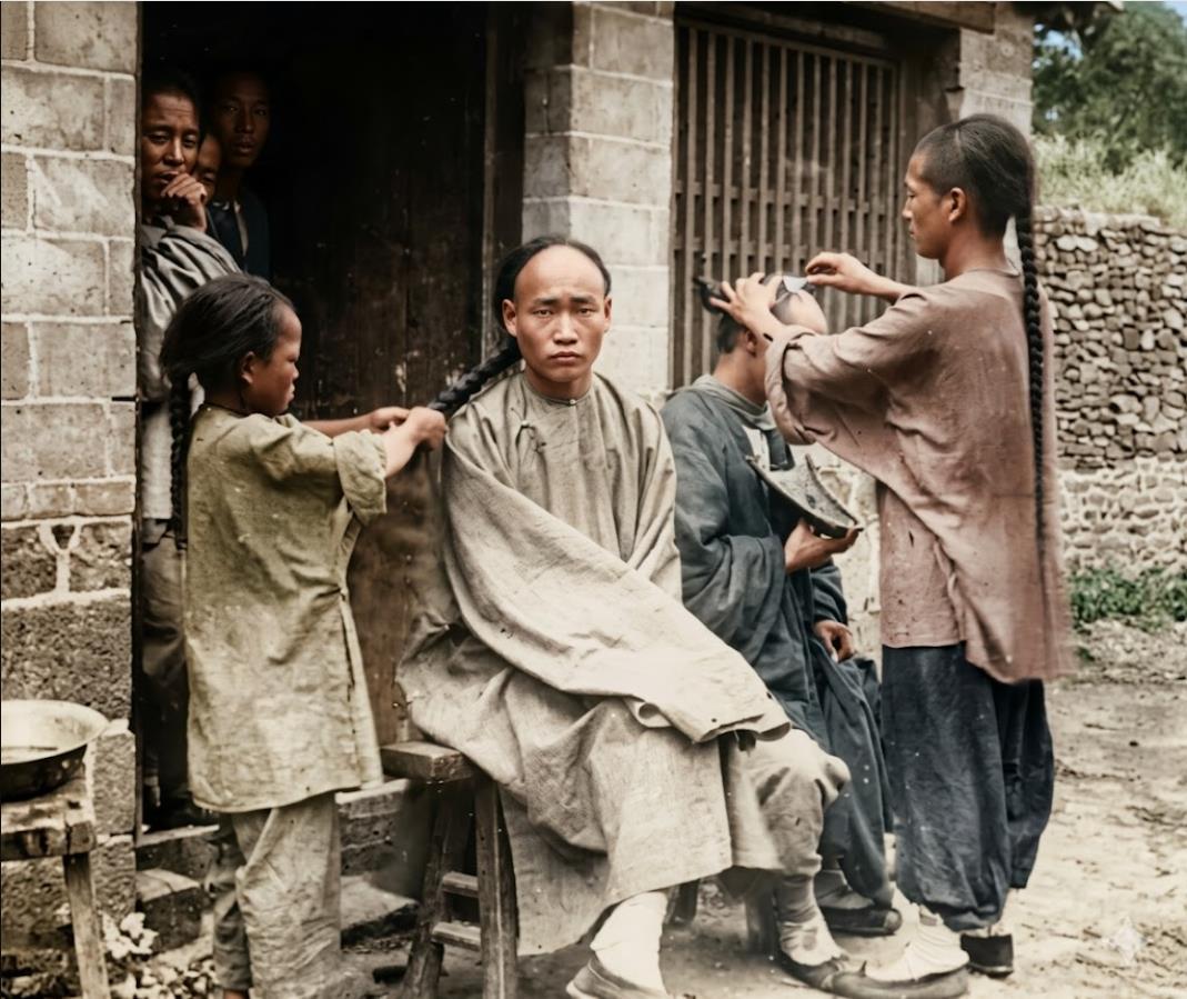 清末，街头剃头摊，剃头师傅正给客人剃前额，他的儿子则在一旁帮客人梳理、编起长长的