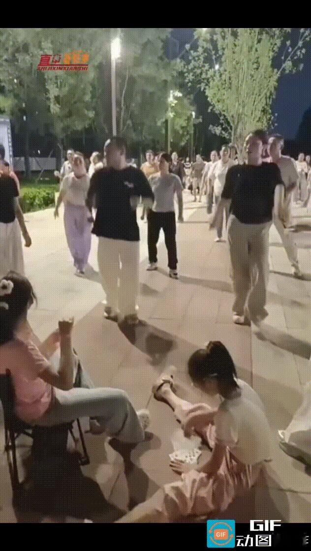 两个女孩坐在广场舞队伍前面玩扑克，一名站在最前方领舞的女子皱着眉头走过来，直接用