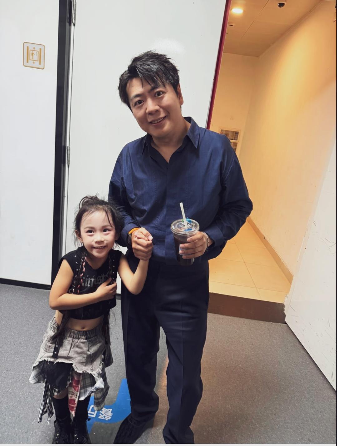 郎朗国际顶尖钢琴演奏家，藏蓝色休闲正装，左手握着一杯冰美式，与小女孩手牵手对着镜