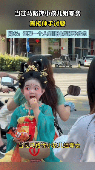一女子在过马路时，瞧见一个身着汉服、打扮精致的小朋友，手里拿着零食。女子没忍住，