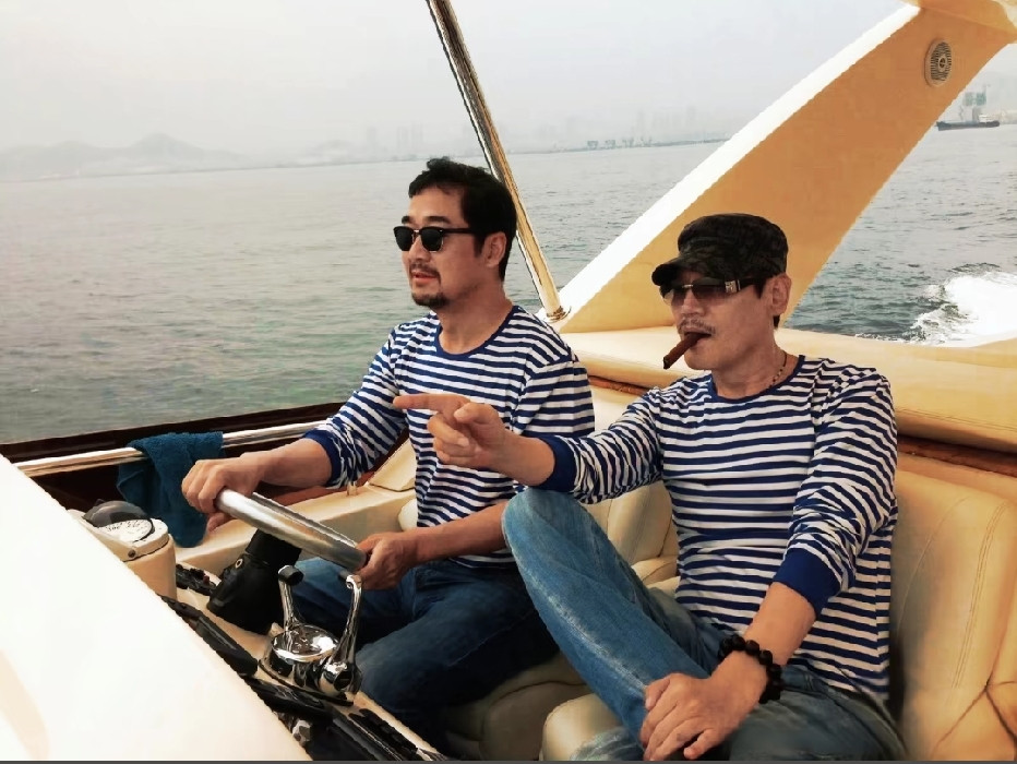 张国立与巍子二人同乘游艇出海休闲时的一幕，两人都穿着海魂衫，张国立手握方向盘，巍子叼着雪茄。