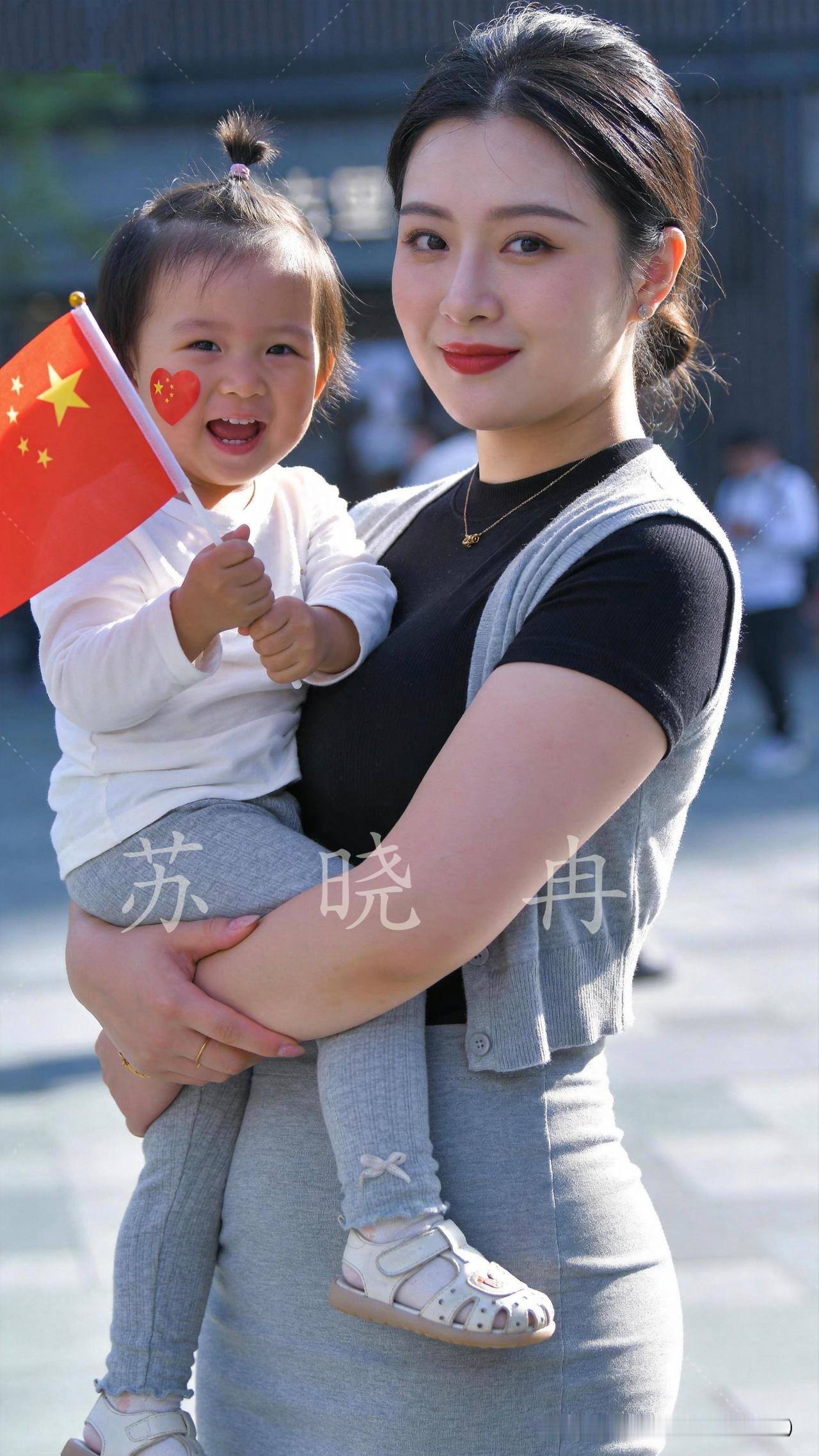 国庆街头喜气洋洋，温柔的妈妈抱着可爱小闺女。小家伙紧紧攥着小红旗，脸颊贴着国旗