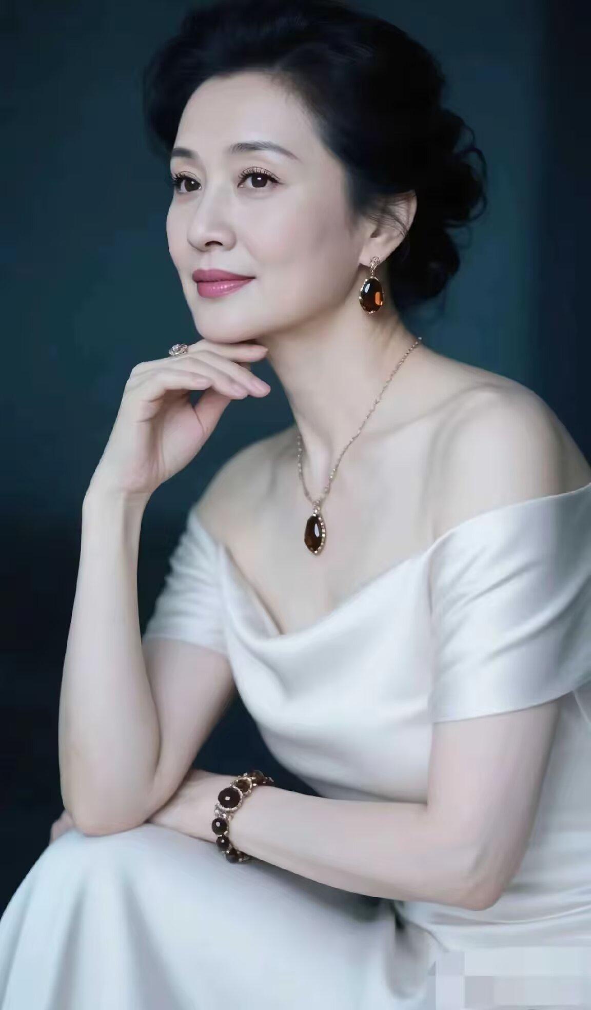 她身着白色露肩礼服，佩戴金色项链与耳环，妆容精致，气质优雅，展现出成熟女性的从容