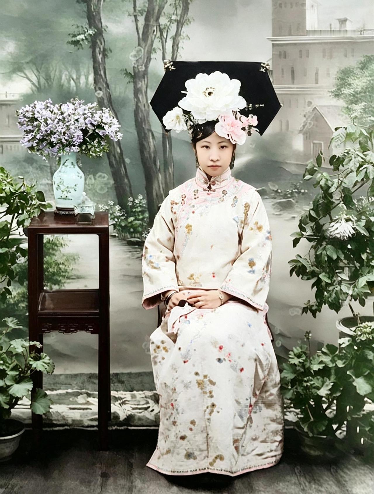 这位女子梳着大拉翅发式，绒绢花点缀其间，一袭旗装华贵考究，面容端丽温婉，举手投足