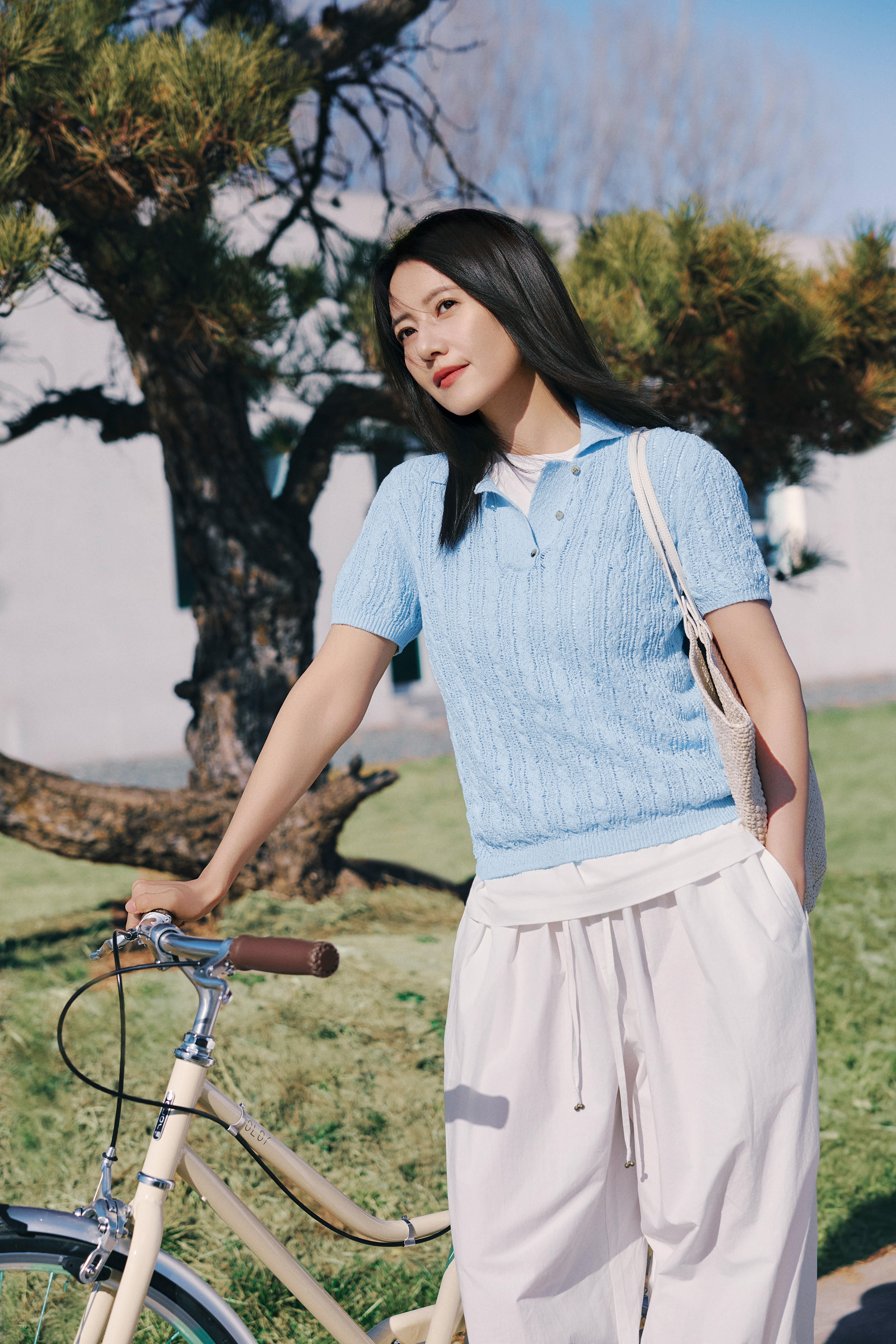 高圆圆站在松枝与暖阳里，浅蓝针织衫衬得眉眼温柔又清亮，黑发被风拂起时，连笑容都