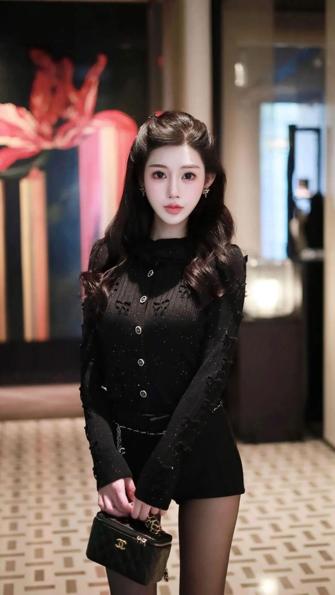 这位美女穿着黑色亮片装，手拿小包，站在画前。紫色亮片裙装亮片裙装