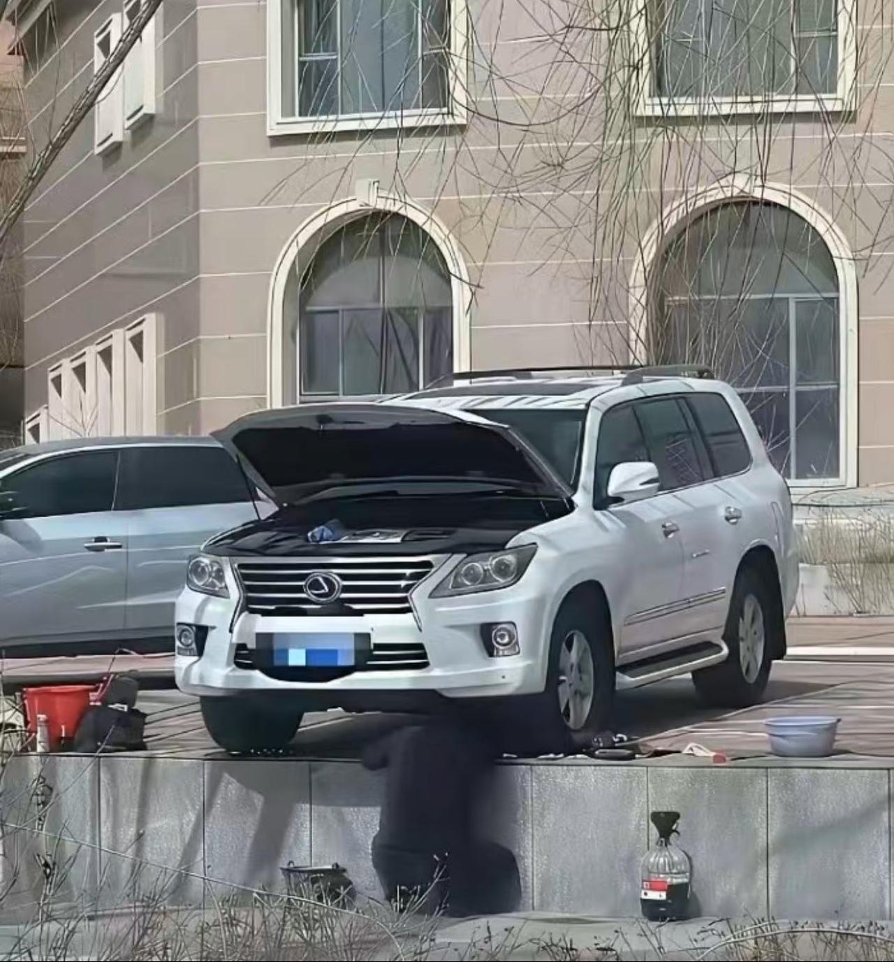 在某居民区内，一辆停放在高凸路缘石上的白色雷克萨斯SUV下方，车主正在亲自动手更