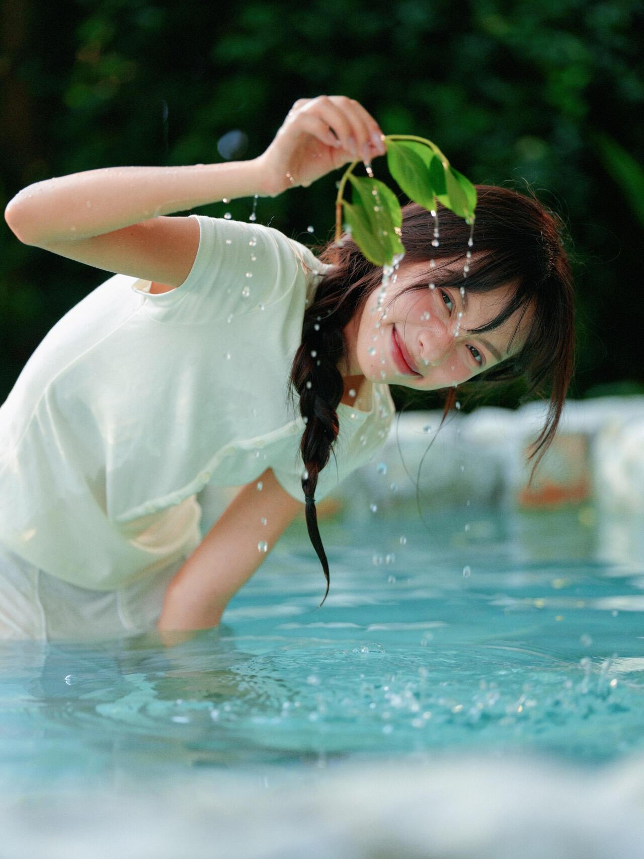 这组写真充满了夏日的清凉感。女孩站在水中，穿着白色的衣服，手里拿着带水的叶子，周