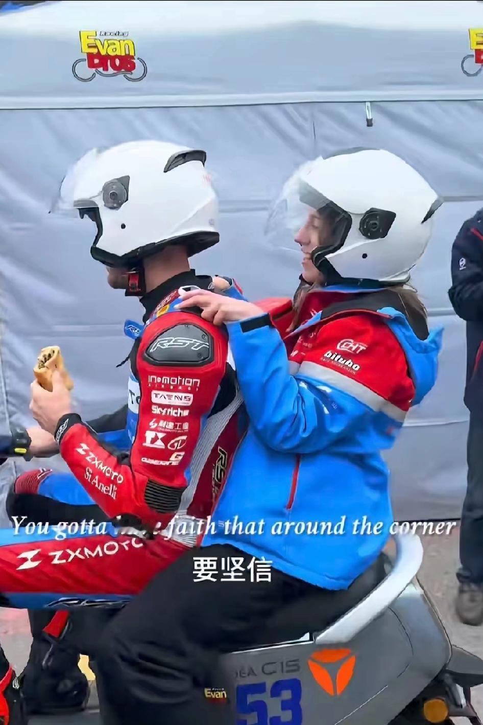 瓦伦丁老婆坐在电瓶车后面，给瓦伦丁捏肩膀放松，骑着雅迪，吃着煎饼果子，挣着光明正