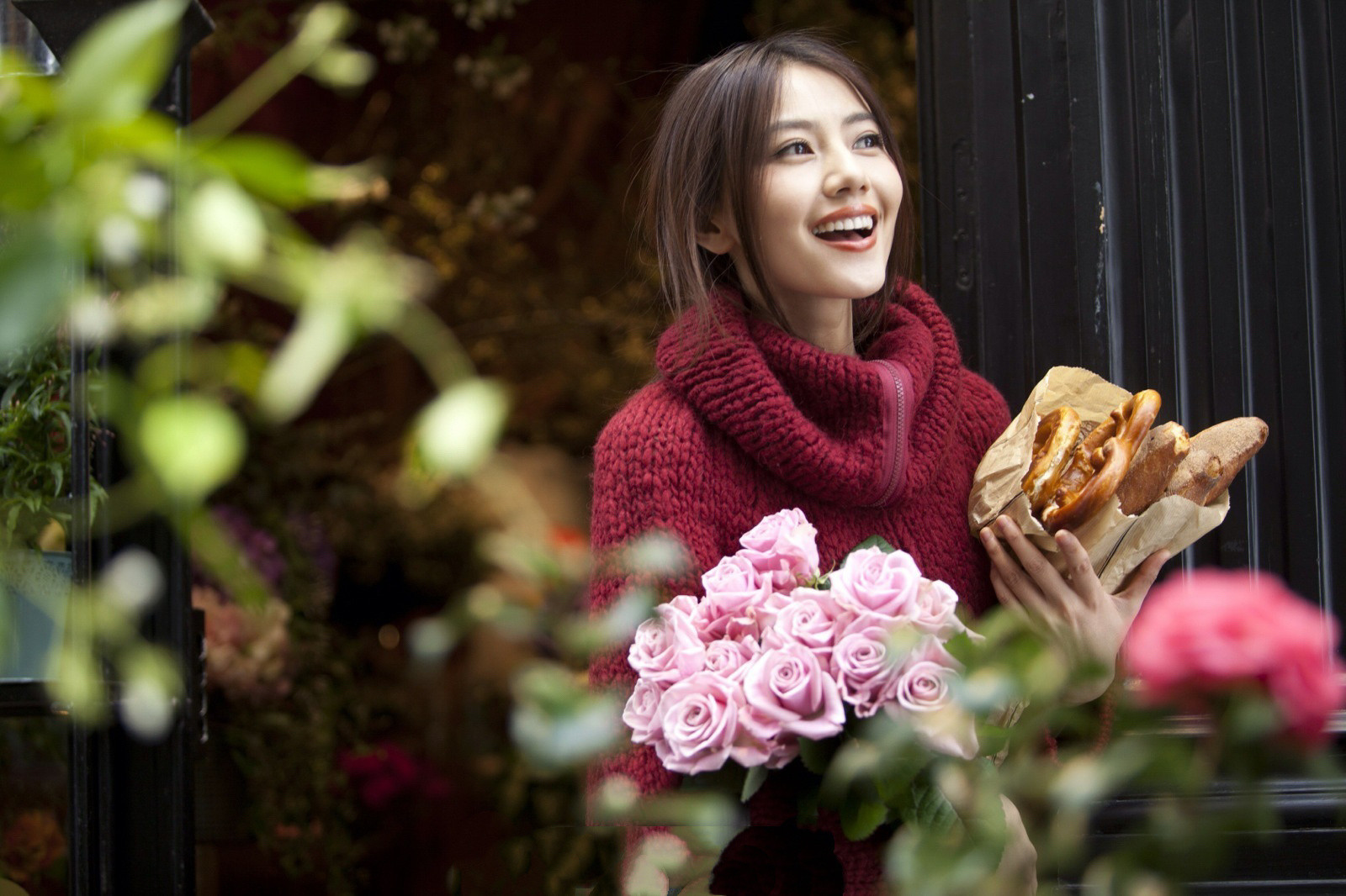 身着红色粗织毛衣，手捧粉玫瑰与法棍面包，笑容明媚又松弛。暖调的红与粉，搭配背景里