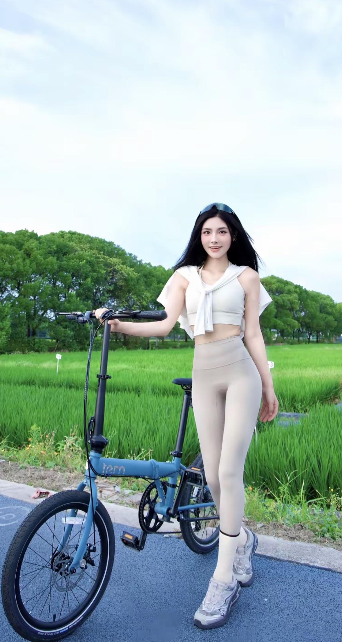 少女身着白色运动装，搭配浅灰紧身裤，倚靠蓝色折叠车驻足绿野小径。微风拂动发丝，笑
