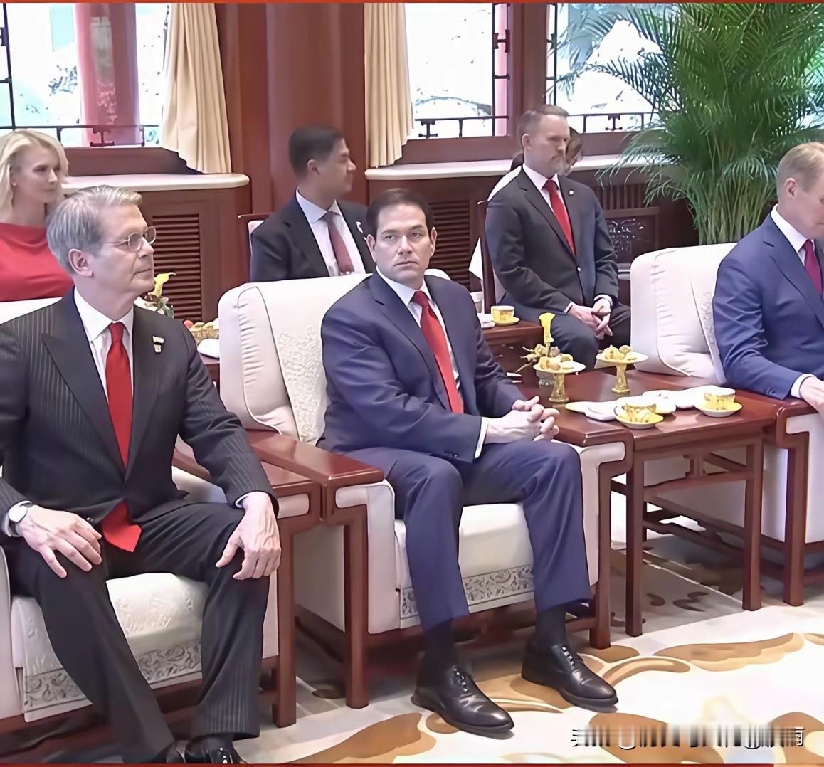 卢比奥在人民大会堂正襟危坐，双手交叉搁腿上，西装皮鞋，打红色领带。人在听讲，眼睛