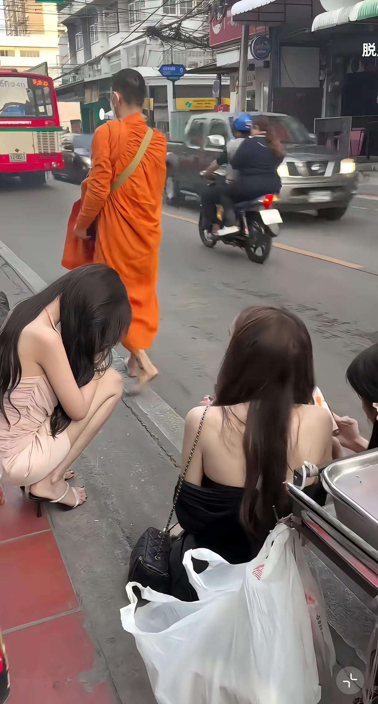 在泰国曼谷街头，几位穿搭时尚的美女正站在一起说笑聊天，一位僧人从旁边走过，这几位