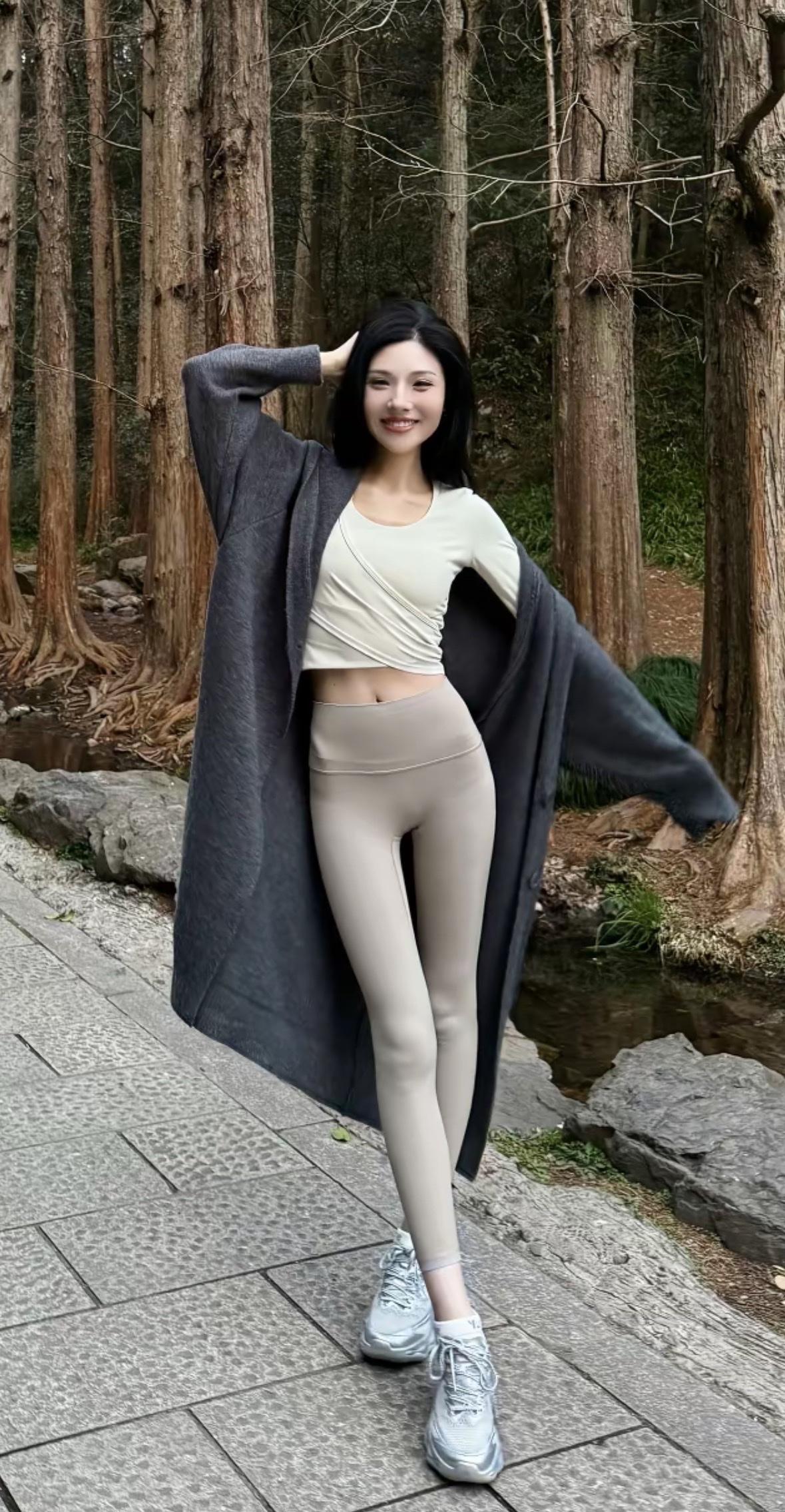 林间小径上，女子身着米色短款上衣与浅灰瑜伽裤，外搭深灰长款开衫，脚踩银白运动鞋。