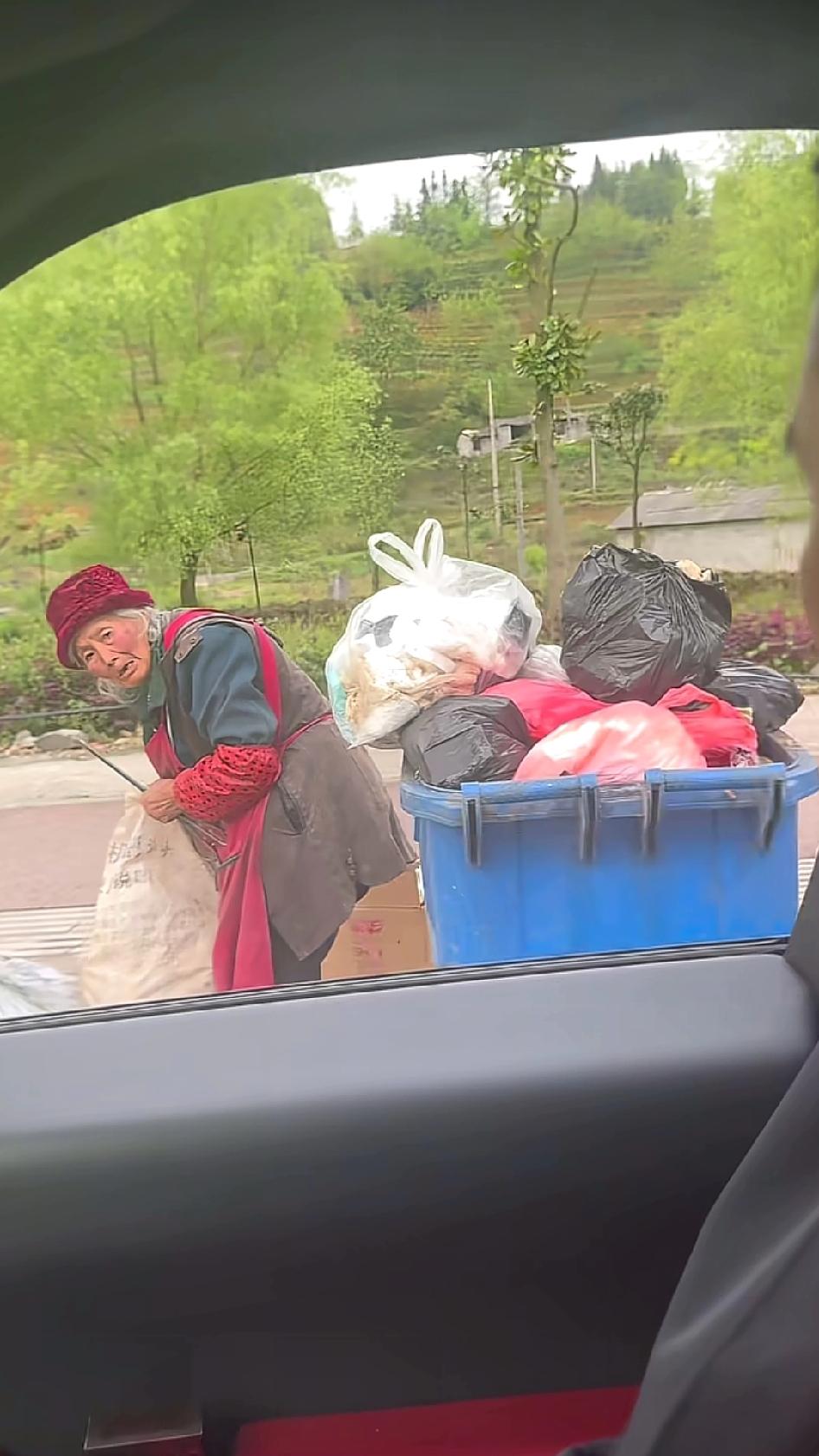 路边一位戴着红帽子，系着红围裙的老奶奶，正在垃圾桶旁收拾废品。一辆轿车停下，车里