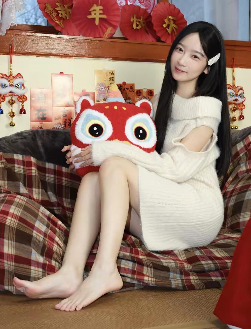 软糯米白毛衣裹着松弛暖意，怀里的虎头玩偶添了几分年味儿。红底金纹的新春装饰错落身