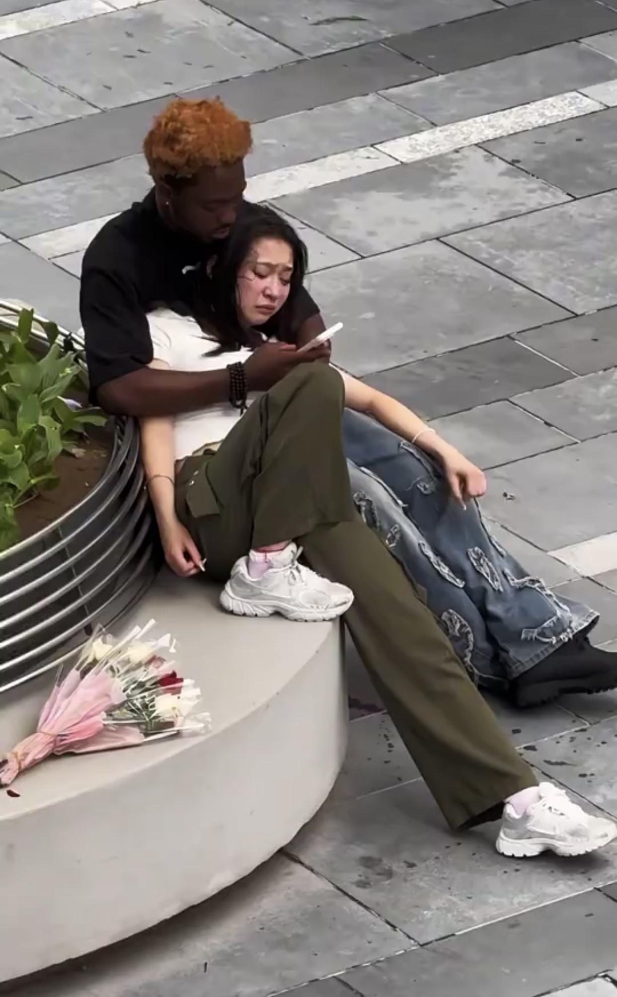 某地大街上，一名女子依靠在一位非洲黑人的怀里痛哭，而黑人则一边搂抱着女子安慰，一