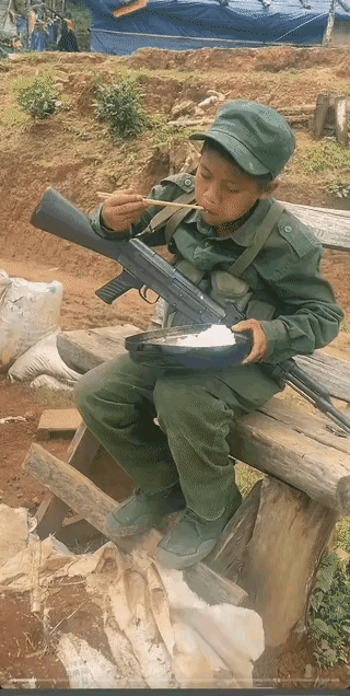 这个小孩儿也就十多岁，穿着军装，背着一支枪，坐在木凳上正在吃饭，用来盛饭的这个东