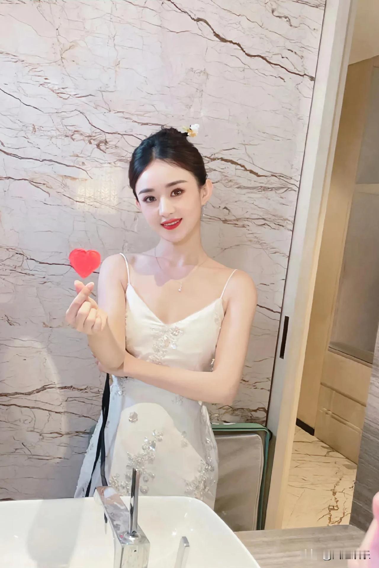 赵丽颖身穿白色吊带，清新脱俗，甜美笑容温暖人心。她轻轻比心，传递爱与正能量，让人