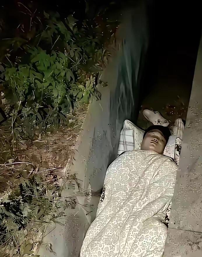 刷到这张图真的瞬间鼻酸！男人裹着花被子，枕着格子枕，就这么睡在路边角落，旁边是杂