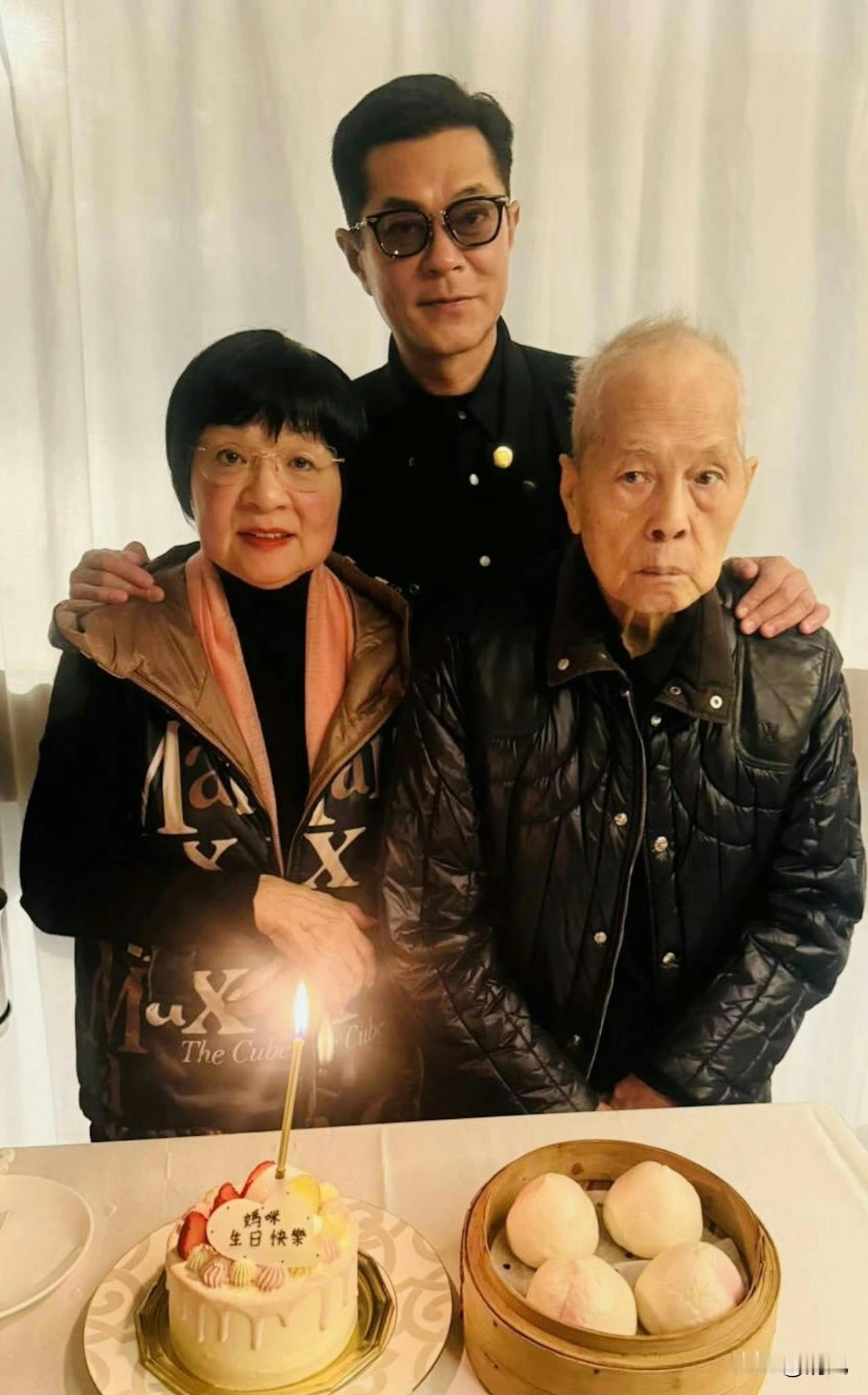 古天乐晒出全家福为妈妈庆生。他身着黑衣，双手搭在父母肩头，桌前摆着定制生日蛋