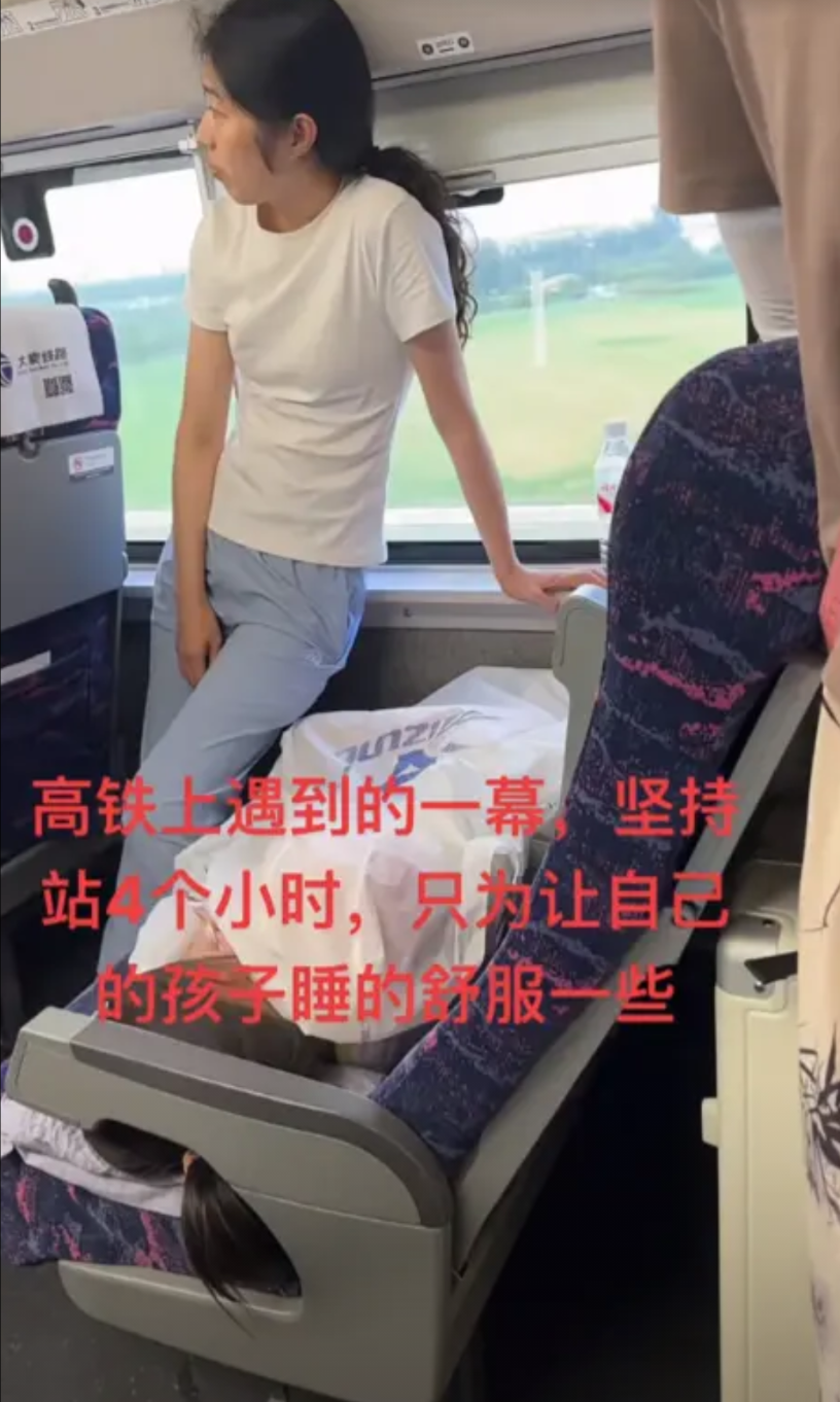 高铁车厢里，一位母亲连续站了四个小时。她面前座位上躺着熟睡的孩子。孩子身下垫