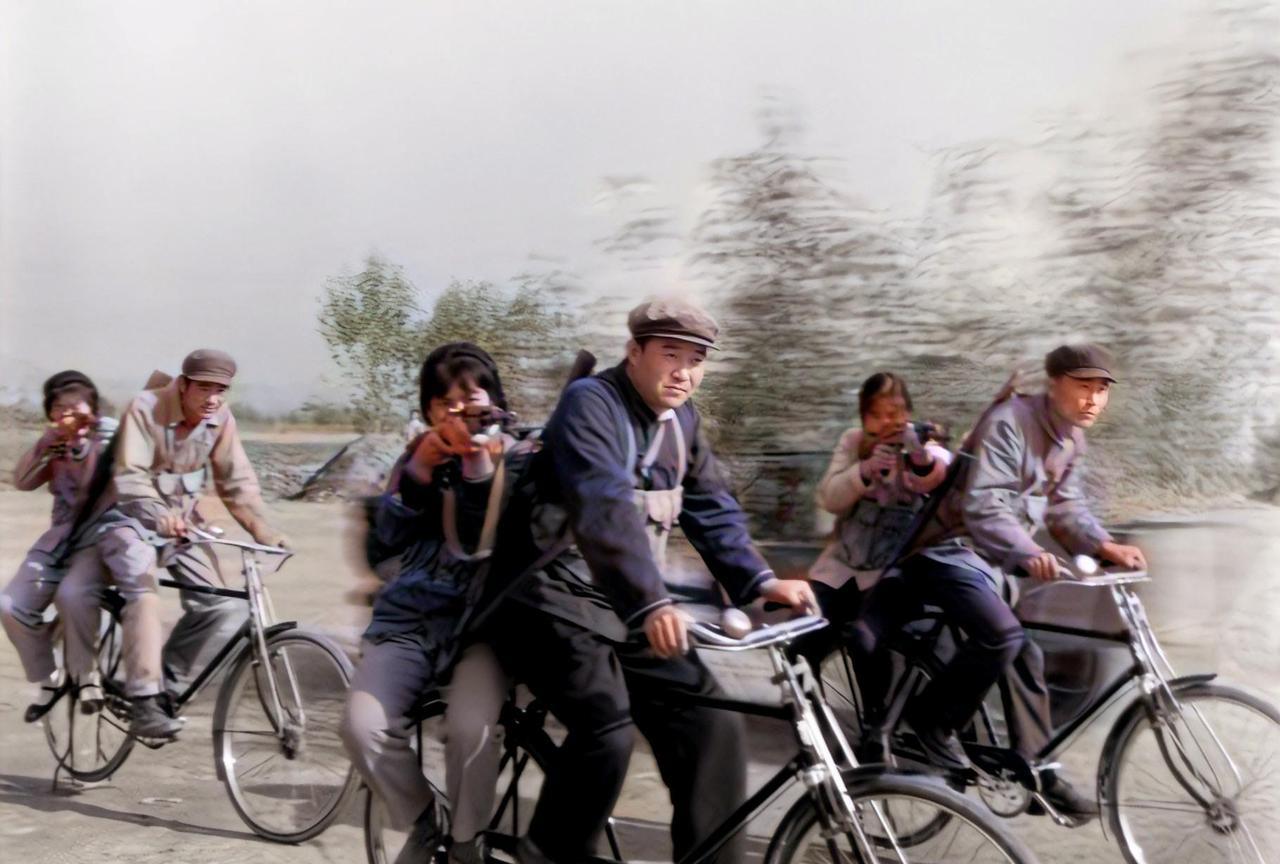 1975年，在厂区外的操场上，3名男民兵奋力蹬着自行车，车后座上的女民兵举枪瞄准