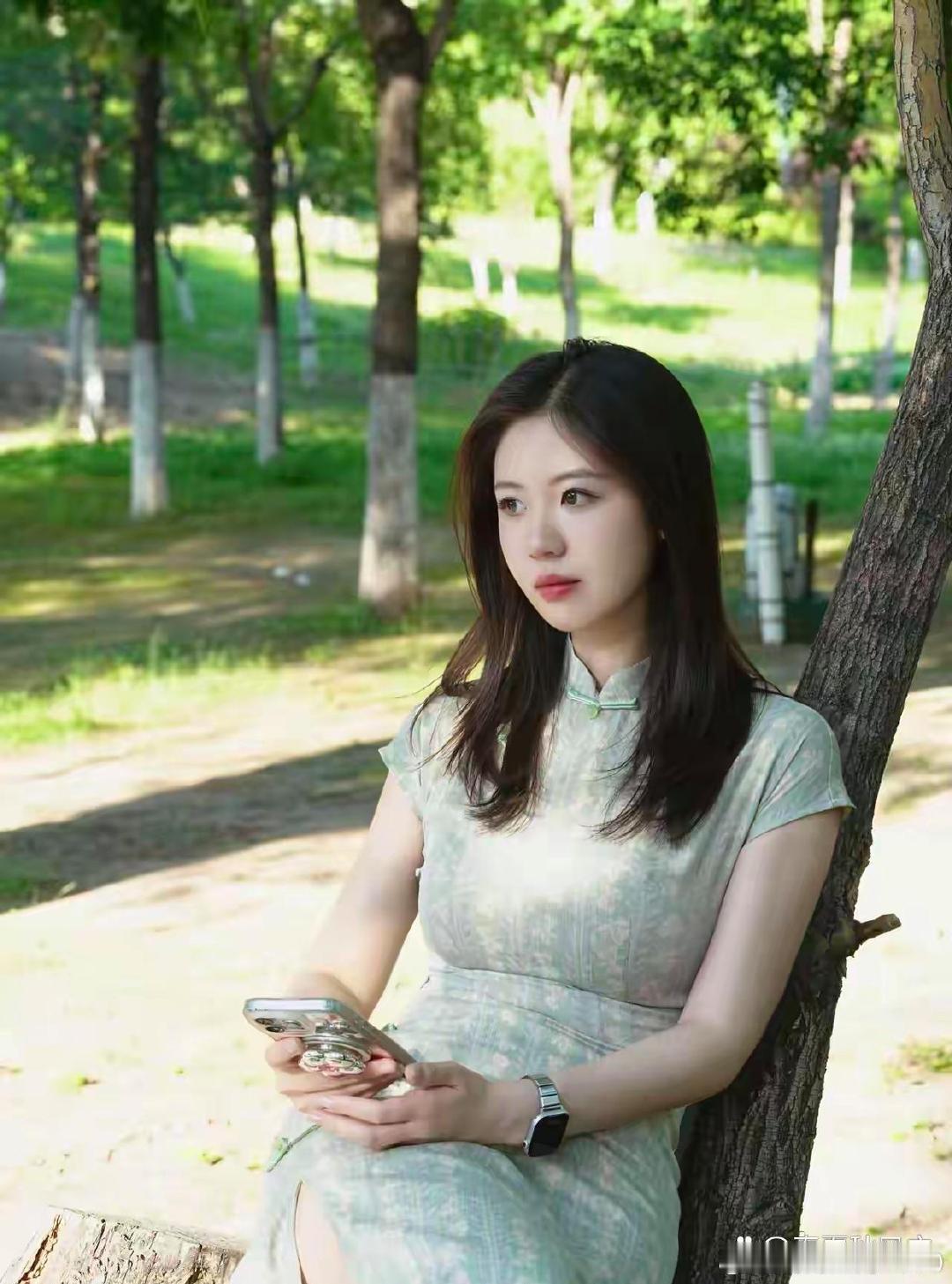 《身着清浅绿暗纹旗袍的女子倚树闲坐，手中握着手机，叶隙筛下的斑驳光影漫在周身，正静静享受这份松弛惬意