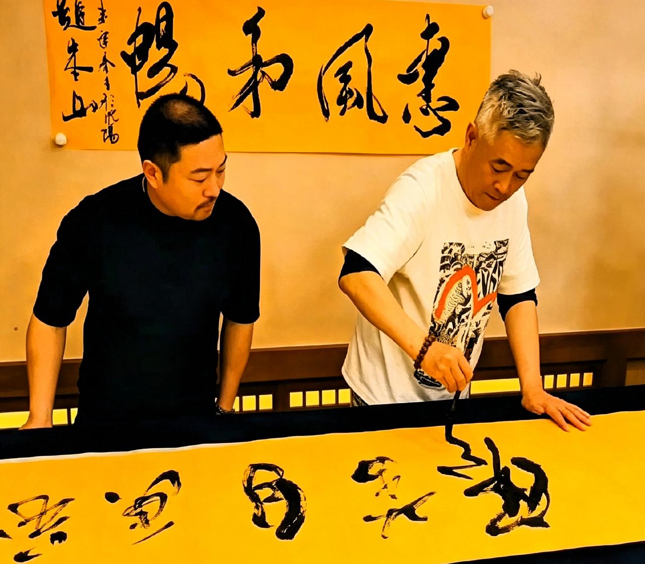本山大叔满头白发，穿着大T恤，正低头写大字呢。旁边哥们儿背着手看得挺认真。这字写