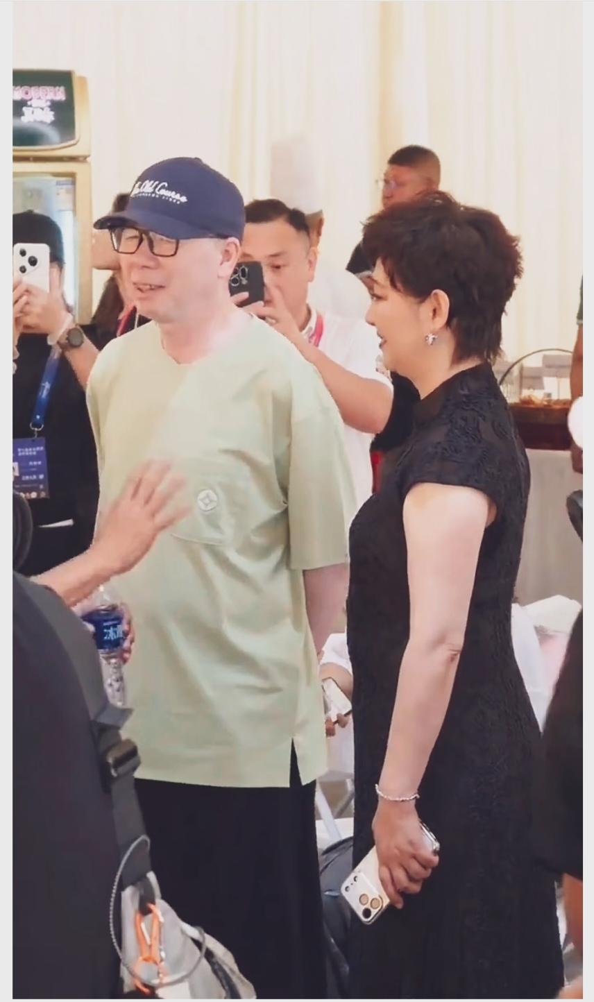 冯小刚导演戴着棒球帽，一身休闲穿搭，皮肤白皙，面带亲切笑容，夫人徐帆站在身边。画