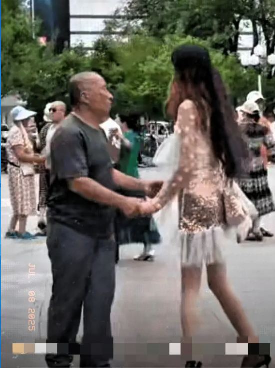 广场上，一位70岁左右的大爷，正在和一位打扮时尚的女子跳舞。从背后看这个女子，一