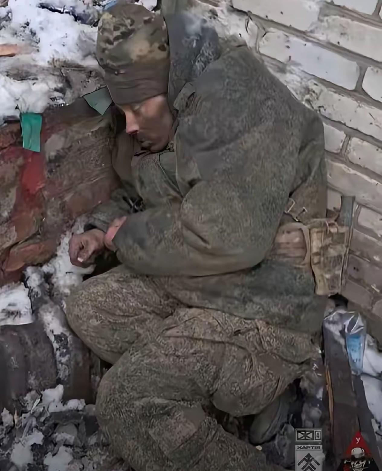 冰天雪地的俄乌战场前线，一名俄军士兵蜷缩在墙角下休息。如此寒冷的天气，穿着迷彩军