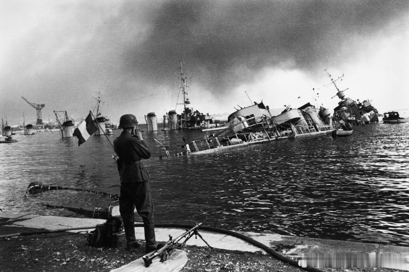 1942年11月27日一名德国机枪手站在土伦港的码头墙上，身后是沉没的法国军舰。