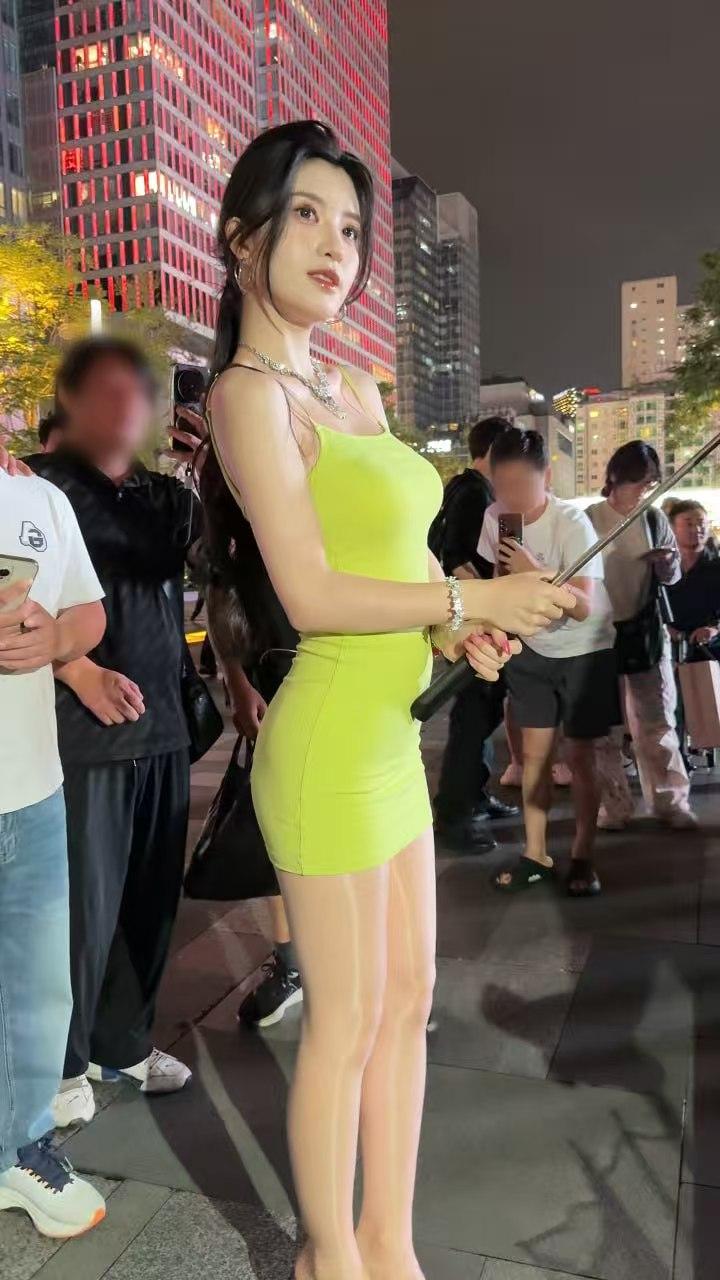 夜色里的商圈广场，亮黄紧身吊带裙裹出窈窕线条，丝袜衬得腿型纤长，她握自拍杆的指尖
