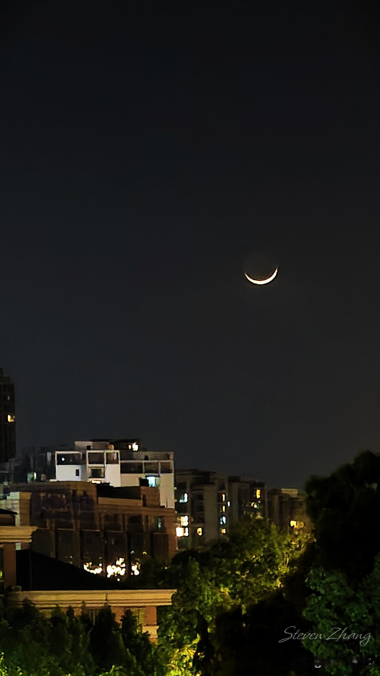 一弯低垂的月，被温柔暮色捧在手心，如同银钩般高挂，枕在万家灯火之上，或许