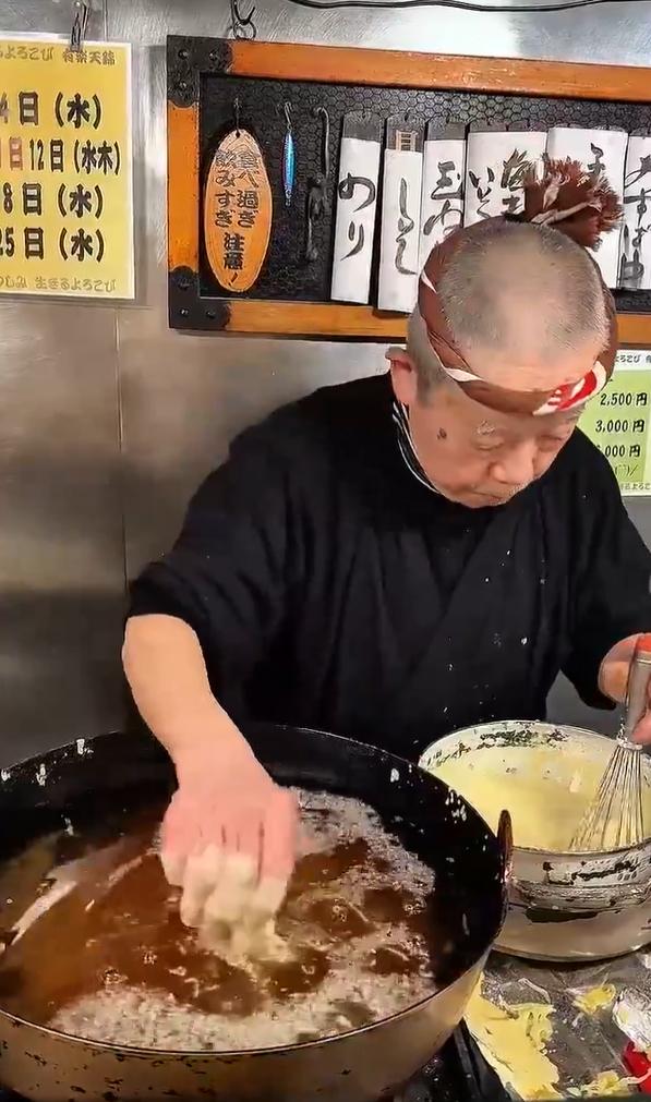 一位年长的日本天妇罗师傅在传统的小店里，徒手将裹好面糊的食材放入油炸锅中，制作盖