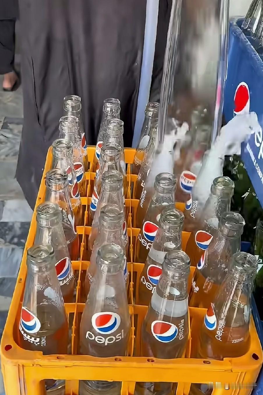 在印度黑作坊内工人把用过的百事可乐瓶子用水管冲干净后，往每个瓶子里放一点黑色的水