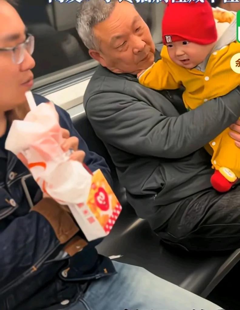 宝宝第一次见奶茶，地铁上爷爷抱着宝宝旁边的一个男子在喝奶茶，宝宝瞪着圆眼，专
