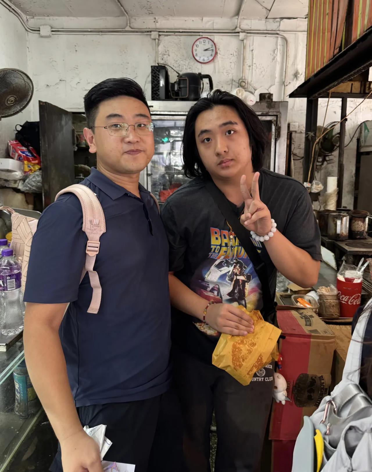 吴镇宇的儿子费曼在香港一家路边摊排队买猪扒包，他虽然只有17岁，但穿衣打扮像一个