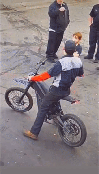 一名男子骑着电摩托正在与人交谈。旁边有个小男孩突然走上前伸手拧了他的车把？电