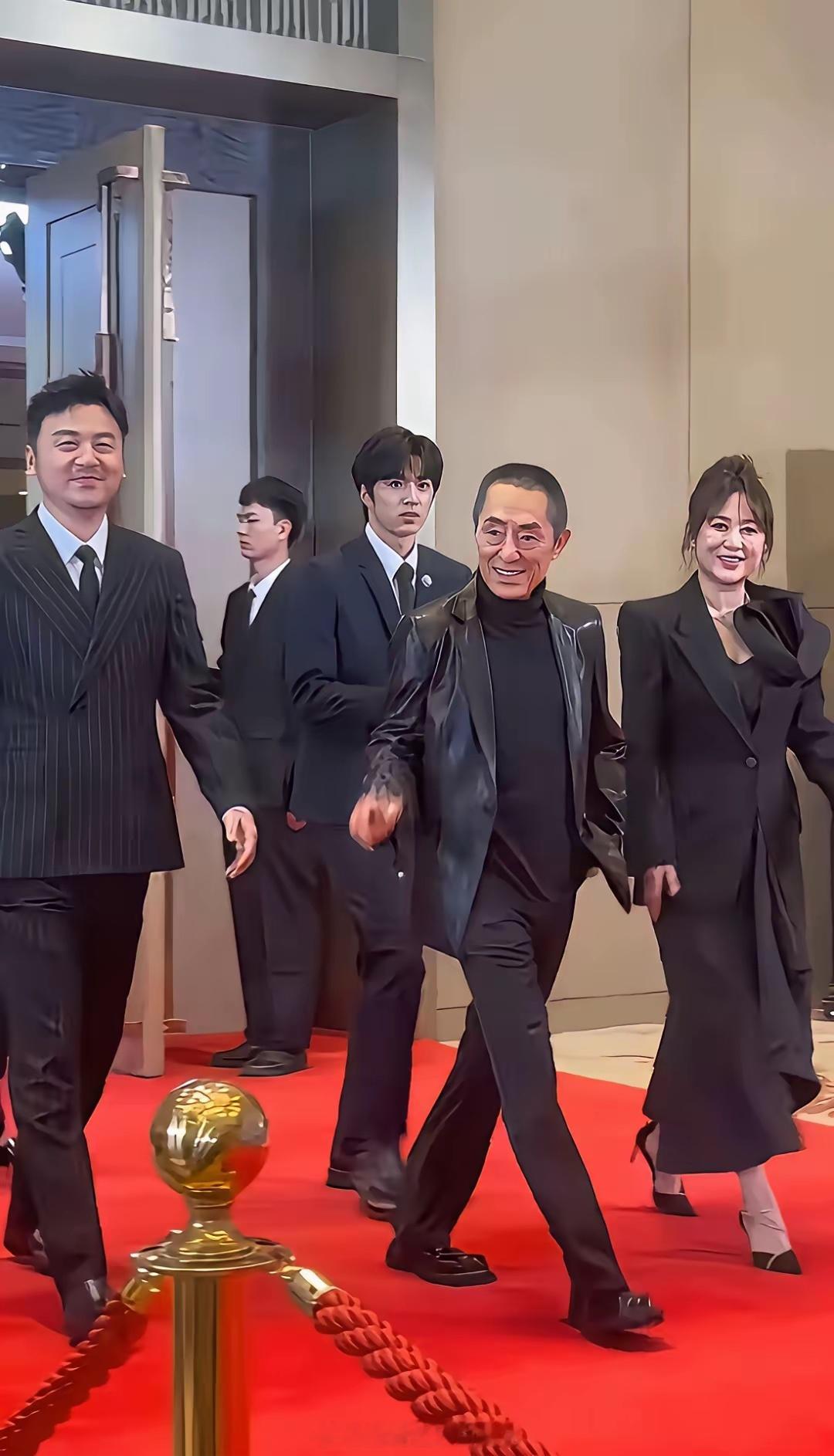 导演张艺谋穿了一件黑色皮衣走在红毯上面，右边是演员雷佳音，左边是演员马丽，张艺谋