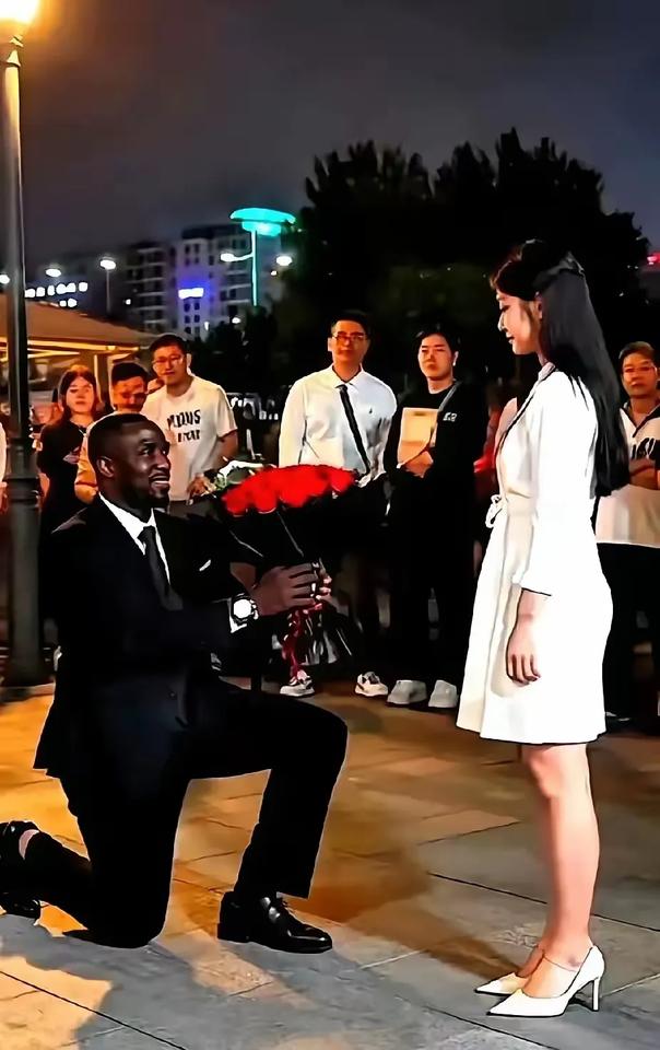 在众目睽睽之下，一西装革履黑人男子捧着玫瑰花，信心满满地微笑着，跪地向一中国姑娘