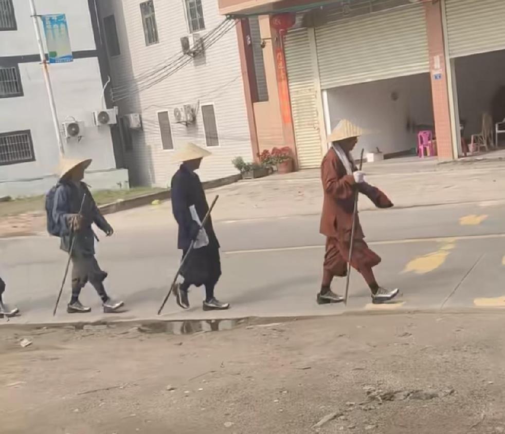 这事就发生在广东梅州，有网友走在路上亲眼撞见。一队人排成一列，拄着棍子，戴着斗
