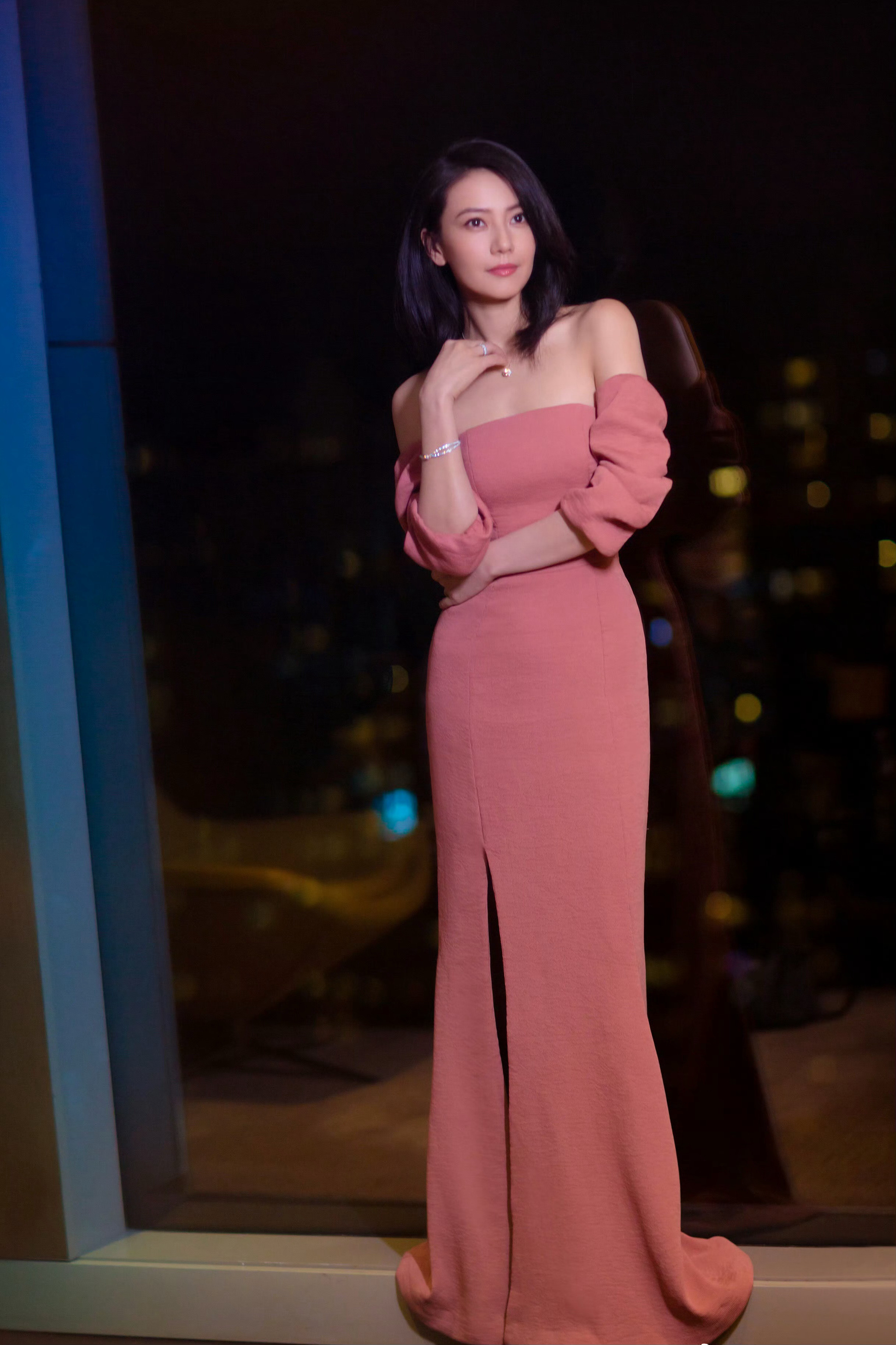 身着豆沙粉露肩长裙，在夜景光影中尽显优雅，锁骨线条精致，钻石配饰闪耀，慵懒卷发更