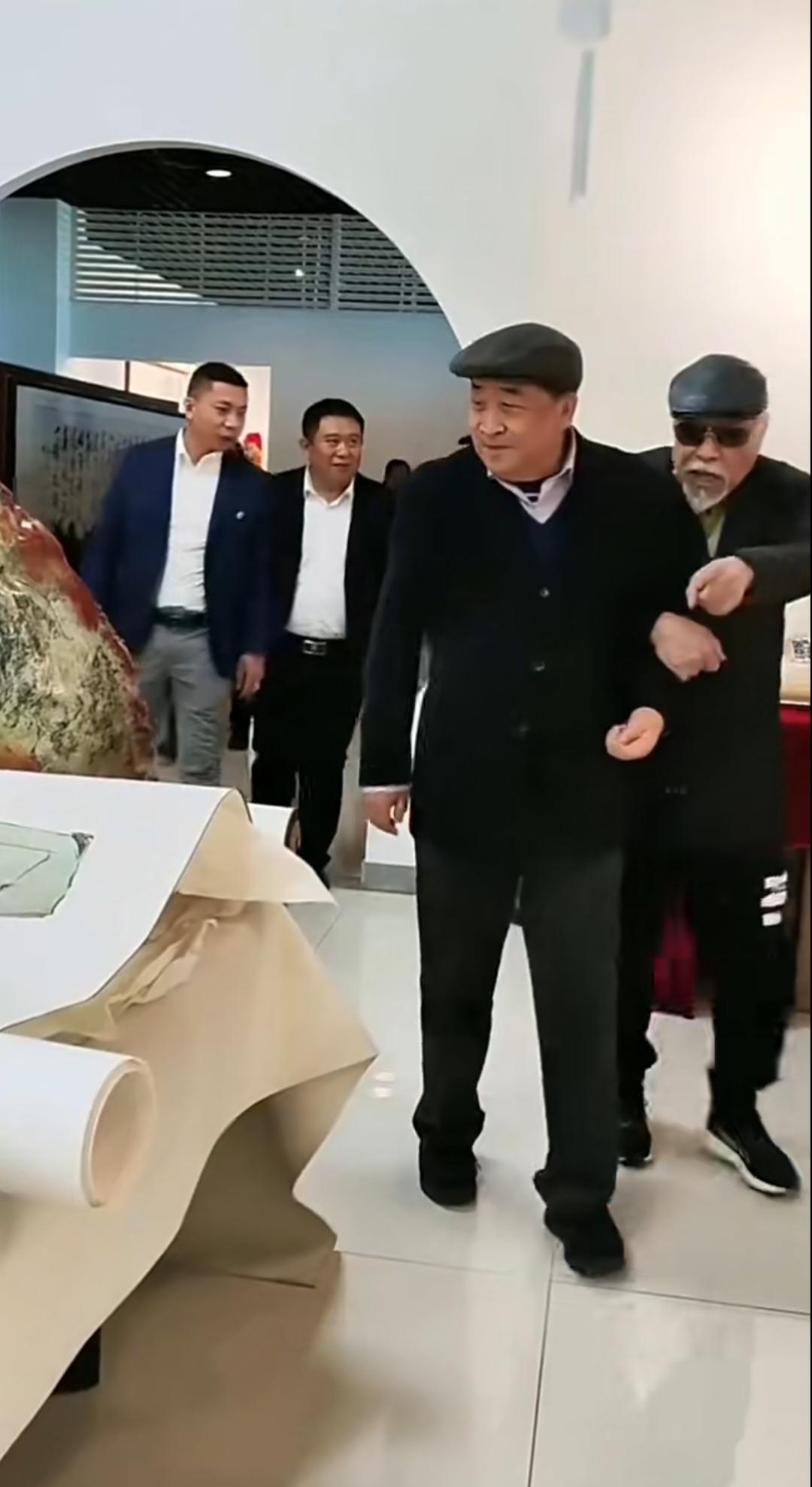 相声泰斗姜昆老师被一位老者挽着胳膊参观画展。戴着鸭舌帽，一身黑色穿搭。尽显艺术家