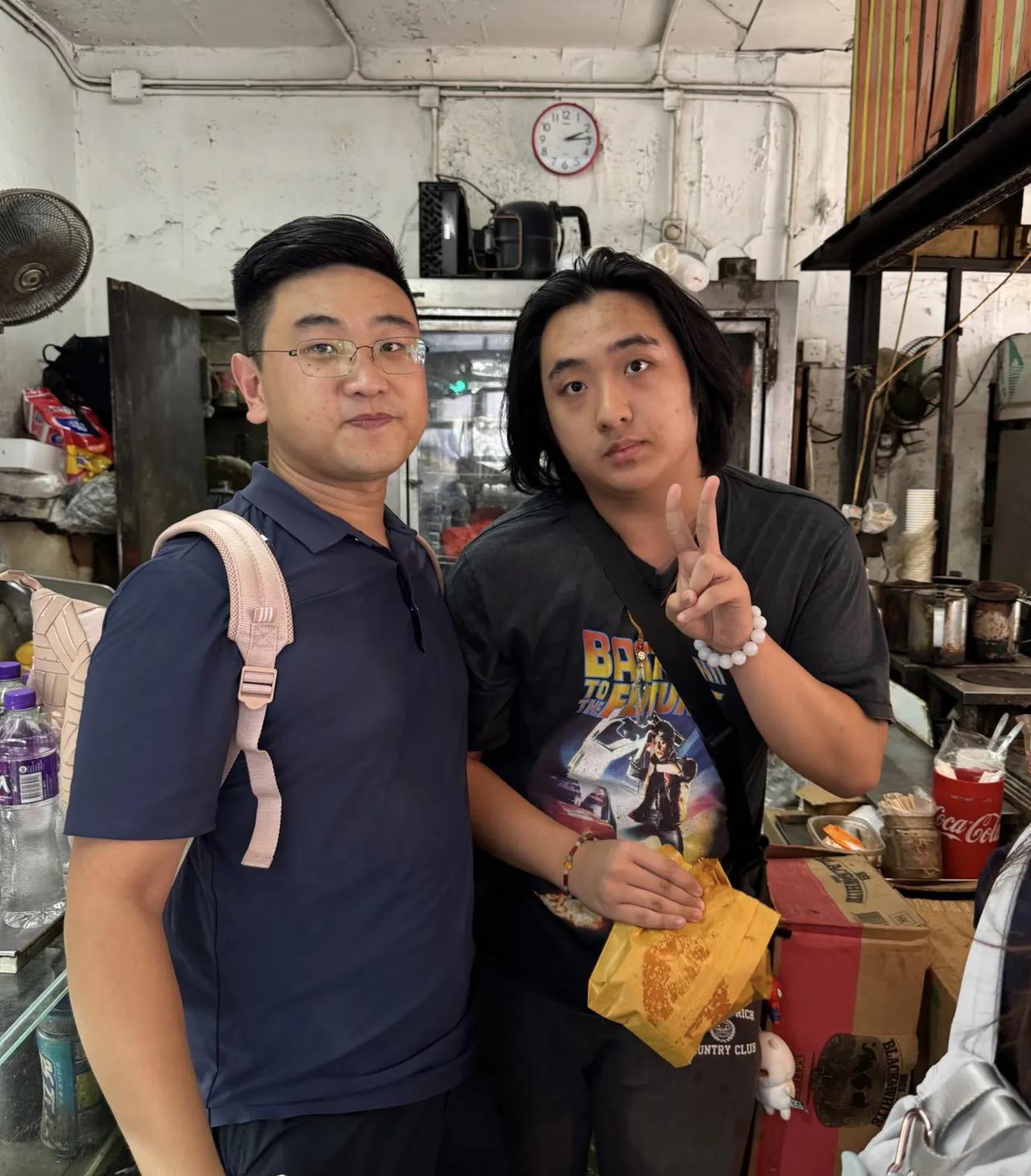 吴镇宇的儿子费曼在香港一家路边摊排队买猪扒包，他虽然只有17岁，但穿衣打扮像一个