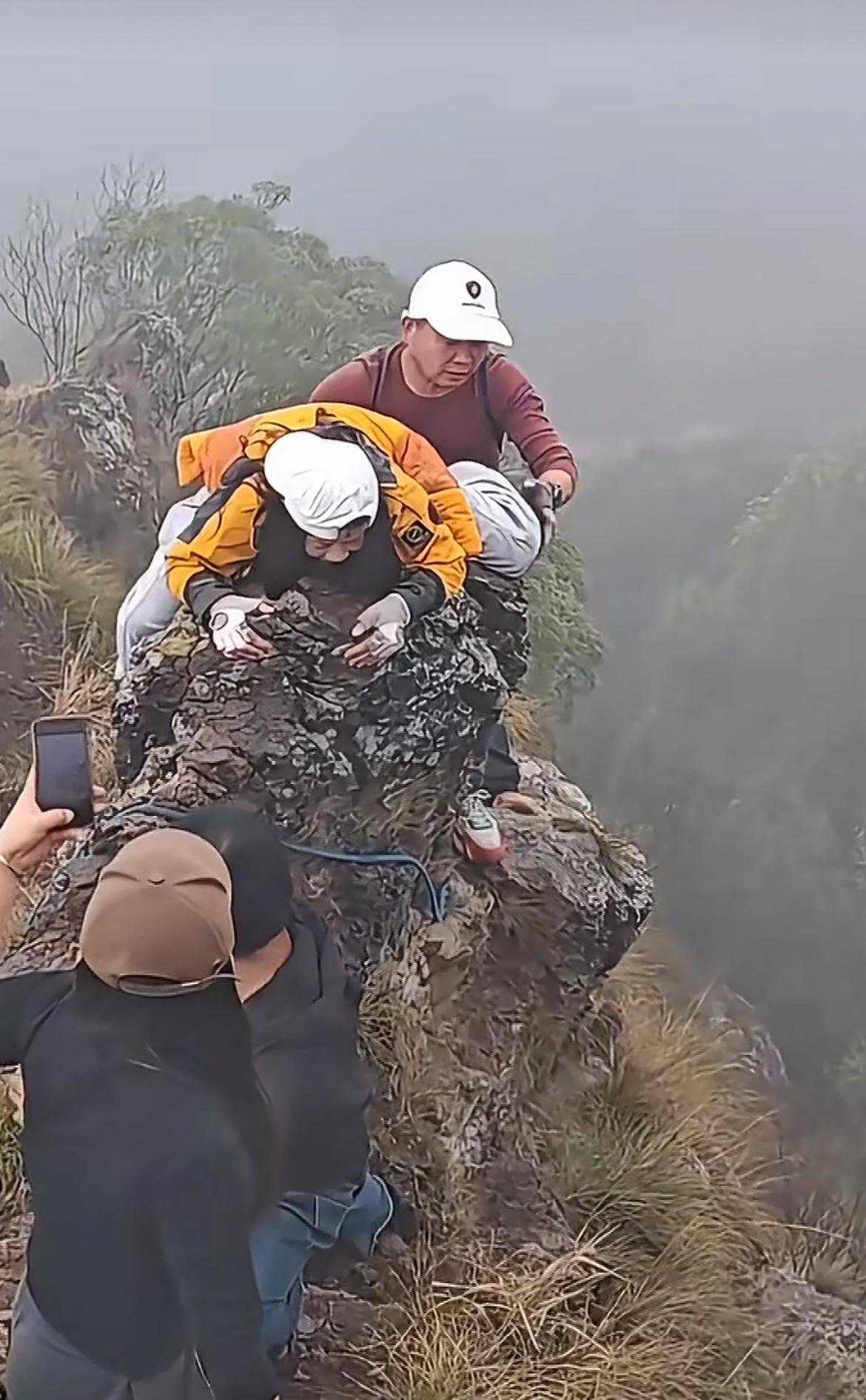 一群驴友爬悬崖，女子吓趴了，趴在石头上崩溃大哭，身后男子不知所措。一群中年男女组