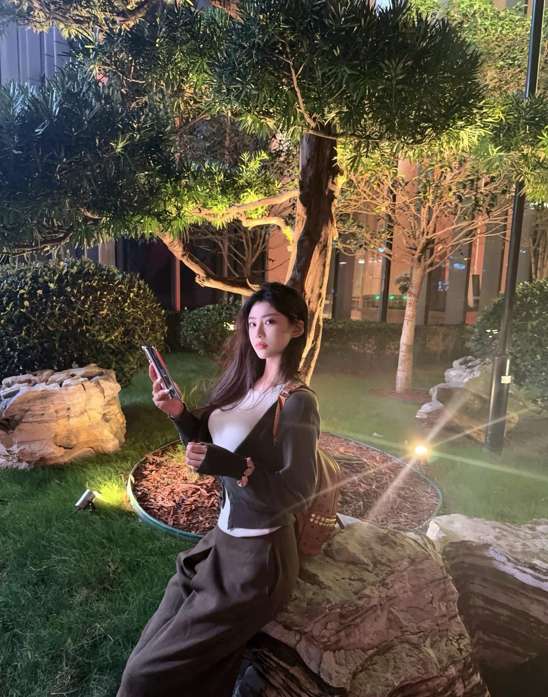 夜晚的公园，静谧而美丽，她坐在石头上，手机照亮了她的脸庞。