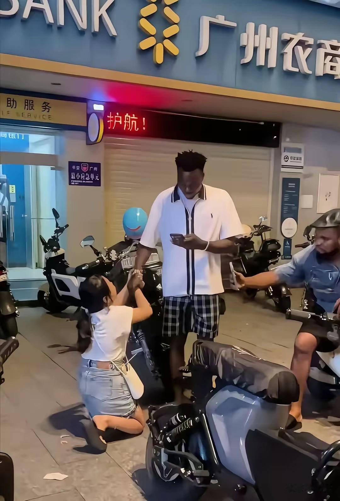 街头一位中国姑娘在地上跪着，对面站着黑人在玩手机。另一位坐在骑电瓶车的黑人则举着