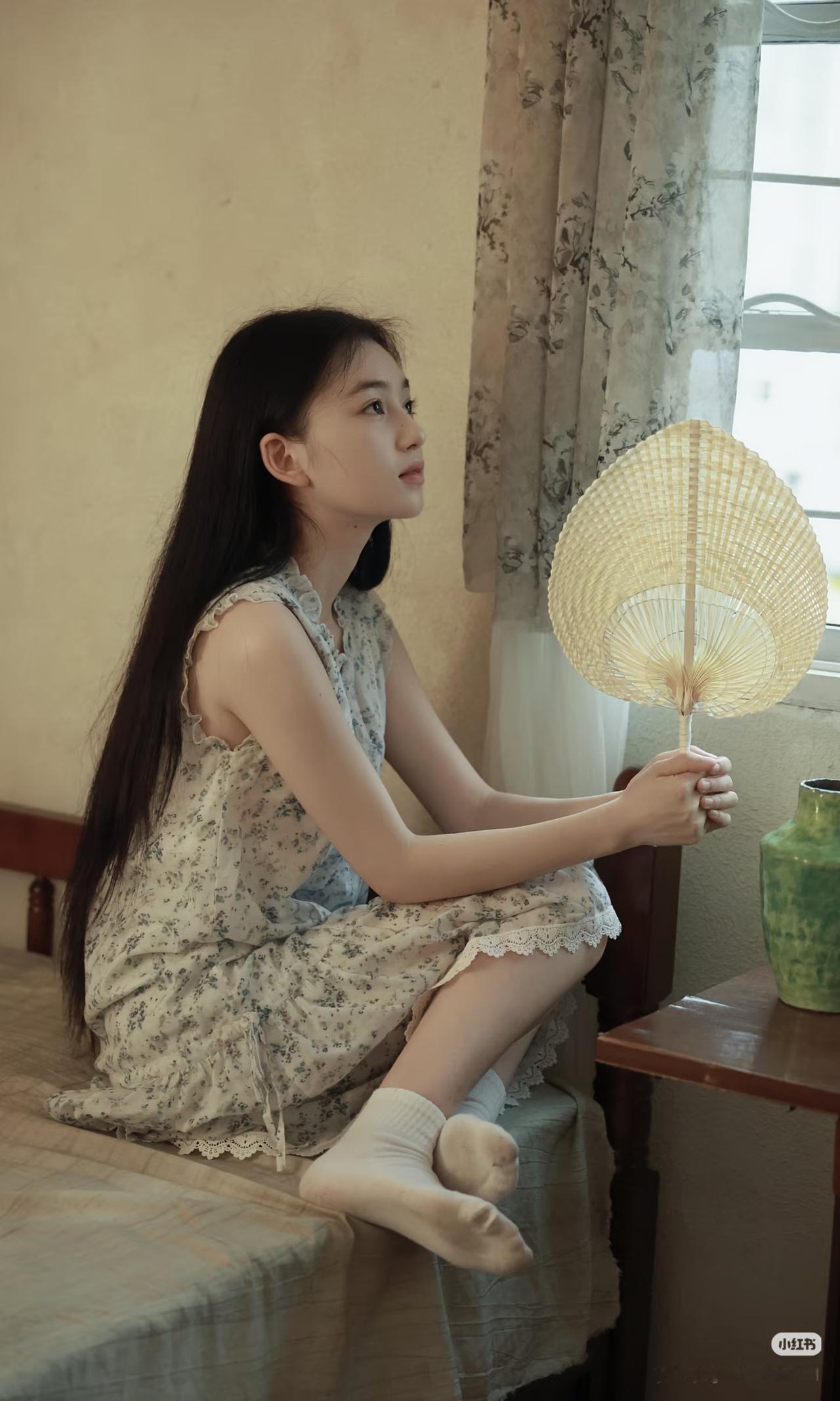 少女静坐窗边，手持团扇，沉思悠远。